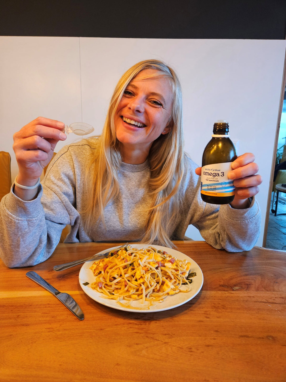 Arianne Meyst aan tafel met een fles Arctic Blue omega 3 full spectrum visolie en een doseerlepel, met een bord pasta voor zich